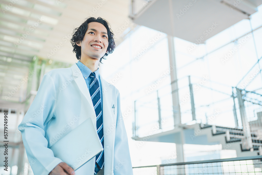 ノートパソコンを持って病院の廊下を歩く白衣を着た若い男性医師 ノートパソコンを持って病院の廊下を歩く白衣を着た若い男性医師