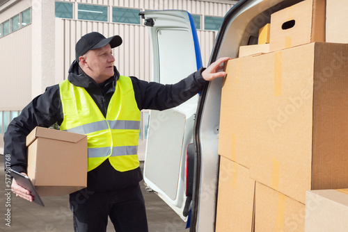 Male courier. Guy takes box from car. Delivery man at work. Courier in reflective vest. Delivery man picks up boxes from warehouse. Courier in industrial zone. Transport company employee