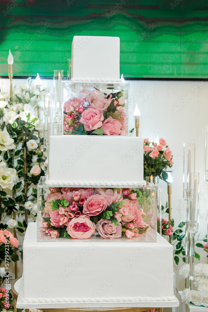 Sweet white engagement cake in banquet hall decorated with artificial flowers and lots of roses behind