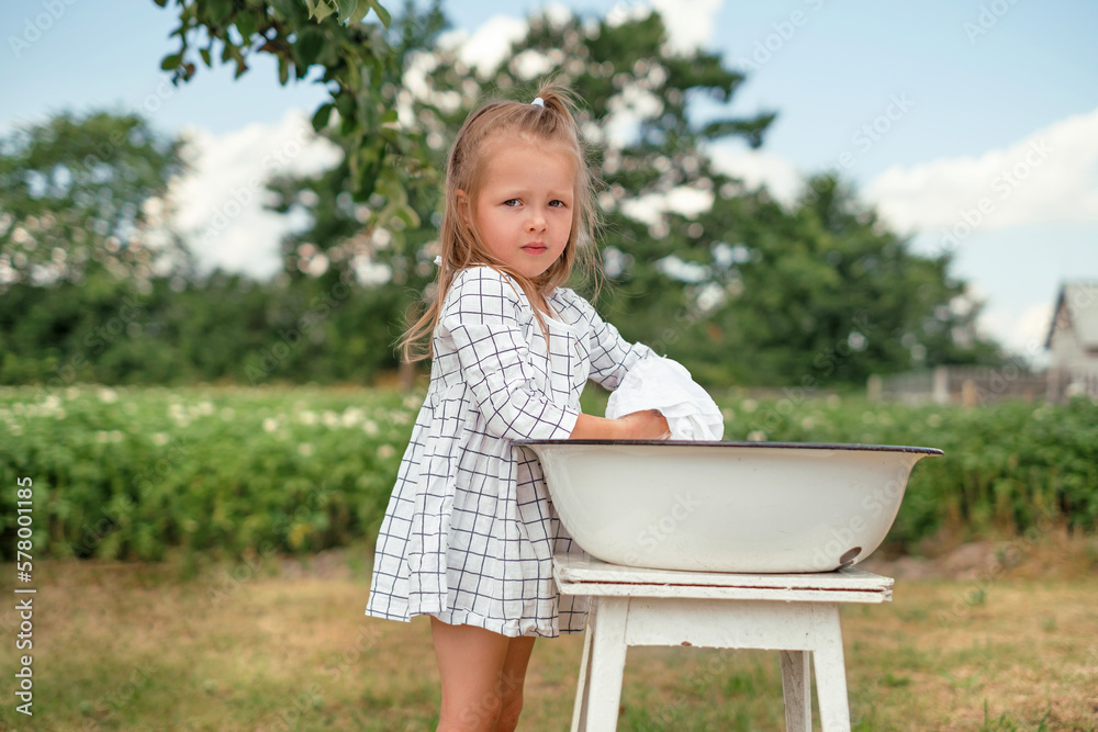 A beautiful attractive girl in a checkered dress is washing clothes in a basin in nature in the garden on a warm summer day. Little mom's assistant in household chores A beautiful attractive girl in a checkered dress is washing clothes in a basin in nature in the garden on a warm summer day. Little mom's assistant in household chores