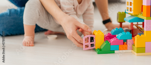 Laughing mother and little daughter playing colorful blocks, constructing tower, sitting on warm floor with underfloor heating, family enjoying leisure time together