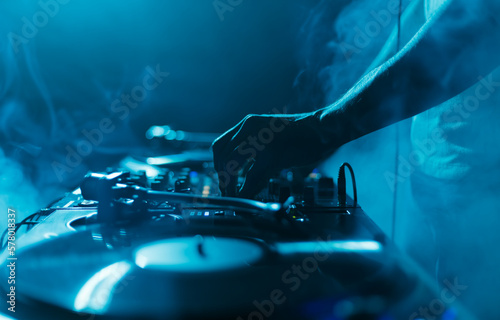 Night club DJ plays set with vinyl records. Close up photo of professional disc jockey mixing music with turntables and sound mixer in smoke and blue lights