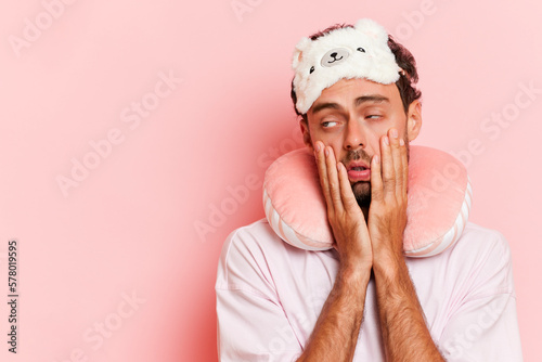 Horizontal shot of tired unshaven caucasian man wears sleeping mask neck pillow looks exhausted while traveling isolated over pink background. Transport, tourism, road trip concept.  photo