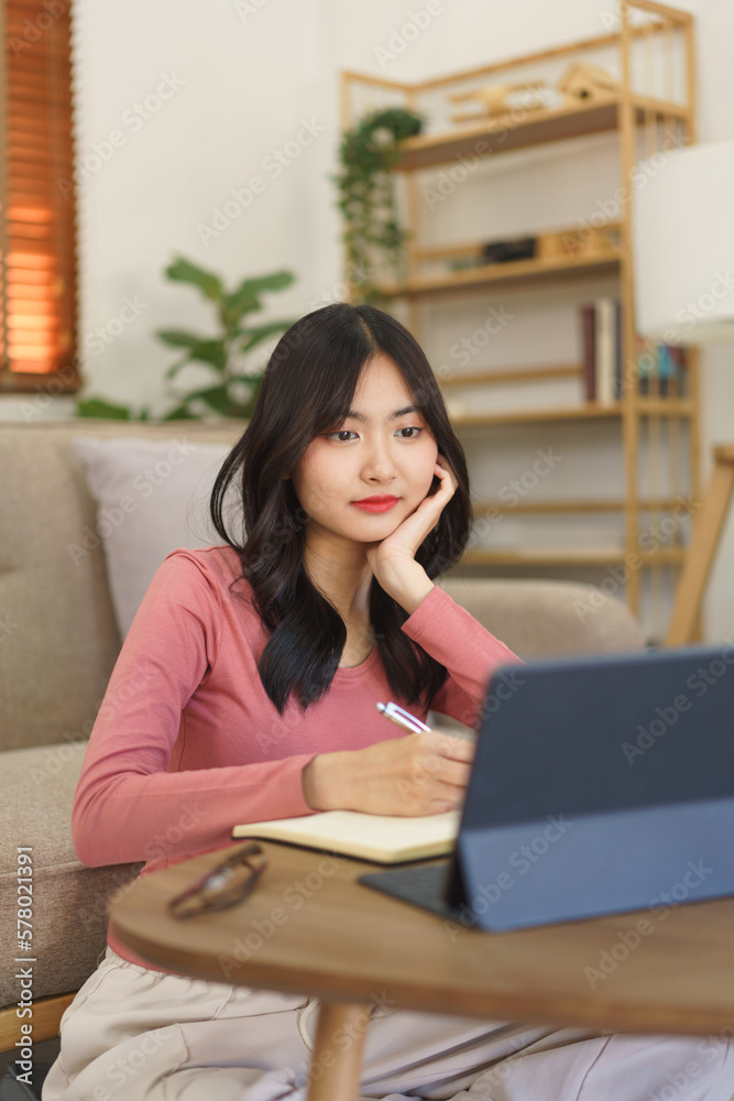 Young businesswoman thoughtful while reading business report on tablet and writing note in notebook Young businesswoman thoughtful while reading business report on tablet and writing note in notebook