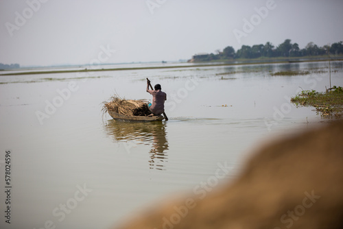 Selective focus on a man rowing a loaded boat to cross the river. Small boat with full of straws carried to another side of the village. Local transportation in remote village during flood.