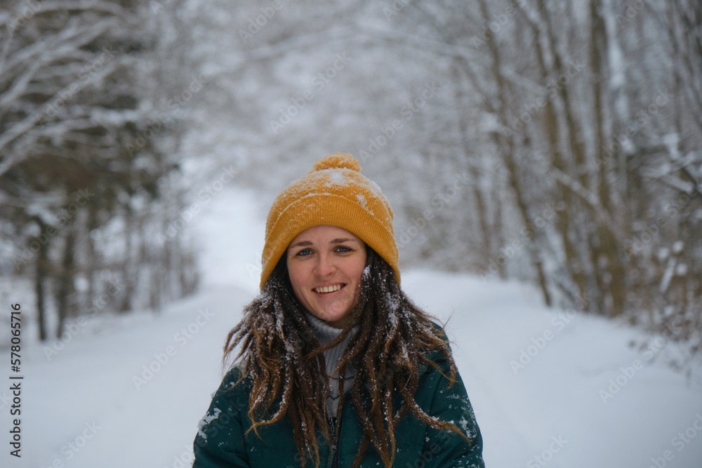 Close up portrait of young smiling pretty Caucasian woman in yellow knitted hat against background of snowy winter forest. Happy European girl with blue eyes and dreadlocks walks alone in winter park. Close up portrait of young smiling pretty Caucasian woman in yellow knitted hat against background of snowy winter forest. Happy European girl with blue eyes and dreadlocks walks alone in winter park.