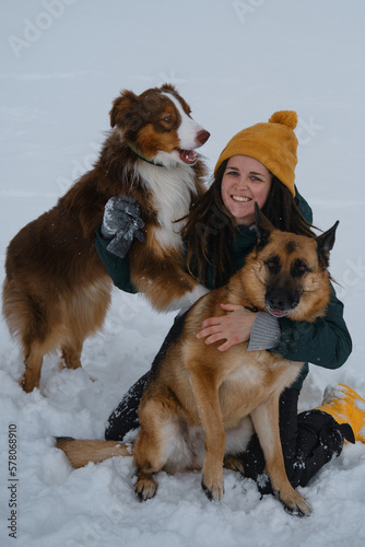 Girl plays with dogs outside in winter. Young pretty Caucasian woman sitting in snow and cuddling with two dogs - German and Australian Shepherds. Concept of warm relationship between owner and pets.
