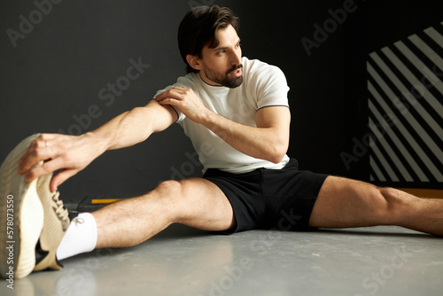 Good-looking brunet bearded guy warming up before sport competition, looking aside with pensive face, having sporty body, sitting on floor with outstretched legs, doing stretching exercises