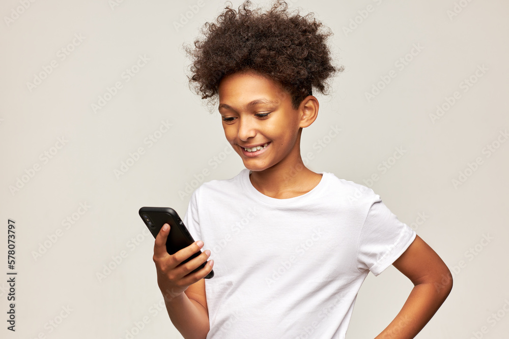 Cute african american boy of 11 using video chat on his mobile phone chatting with his girl mate, smiling widely, feeling happy and in love, isolated on gray studio background in white mockup t-shirt Cute african american boy of 11 using video chat on his mobile phone chatting with his girl mate, smiling widely, feeling happy and in love, isolated on gray studio background in white mockup t-shirt