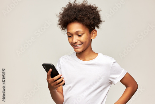 Cute african american boy of 11 using video chat on his mobile phone chatting with his girl mate, smiling widely, feeling happy and in love, isolated on gray studio background in white mockup t-shirt