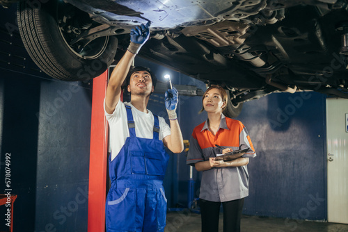 Asian repairman or car mechanic showing and explaining vehicle broken part and maintenance report to female office worker at auto repair shop. After service concept