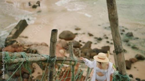 Young girl in swimsuit, white shirt and straw hat walks up a rocky hill stot from above. Blonde female going up the stairs cut in red rock. Young caucasian woman climbs scenic hill using wooden stairs photo