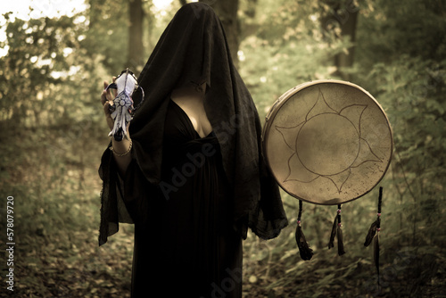 Young witch in twilight forest holding shamanic drum and wolf skull. A mysterious pagan priestess with covered face. Mystic Halloween witchy atmosphere. Occulture, magic, spiritual, witchcraft. photo