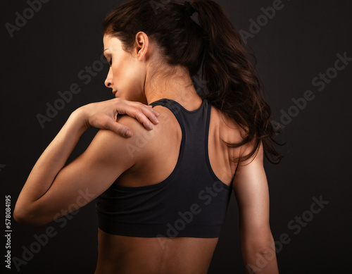 Female sporty muscular with ponytail doing stretching workout of the shoulders, blades in sport bra, holing the neck the hand on dark background with empty copy space. Back view.