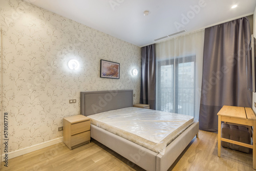 Interior of the bedroom with a large bed  nightstand and window. .Light wall decor and wood flooring.