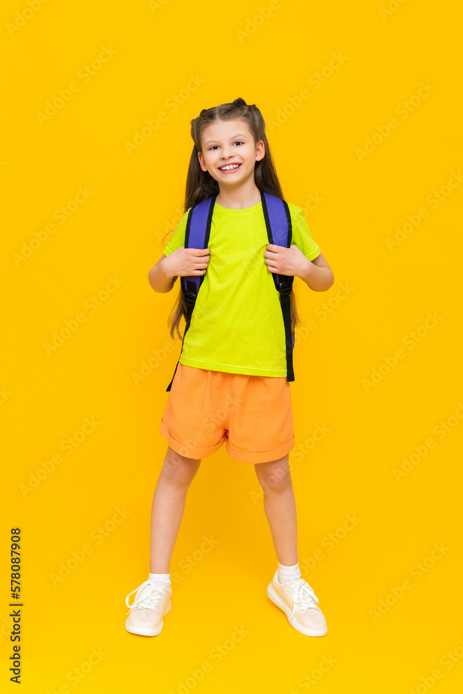 A child with a school backpack. A beautiful little girl in full height, in shorts and a T-shirt. Education during the summer holidays. Preparatory courses for children. Yellow isolated background. A child with a school backpack. A beautiful little girl in full height, in shorts and a T-shirt. Education during the summer holidays. Preparatory courses for children. Yellow isolated background.