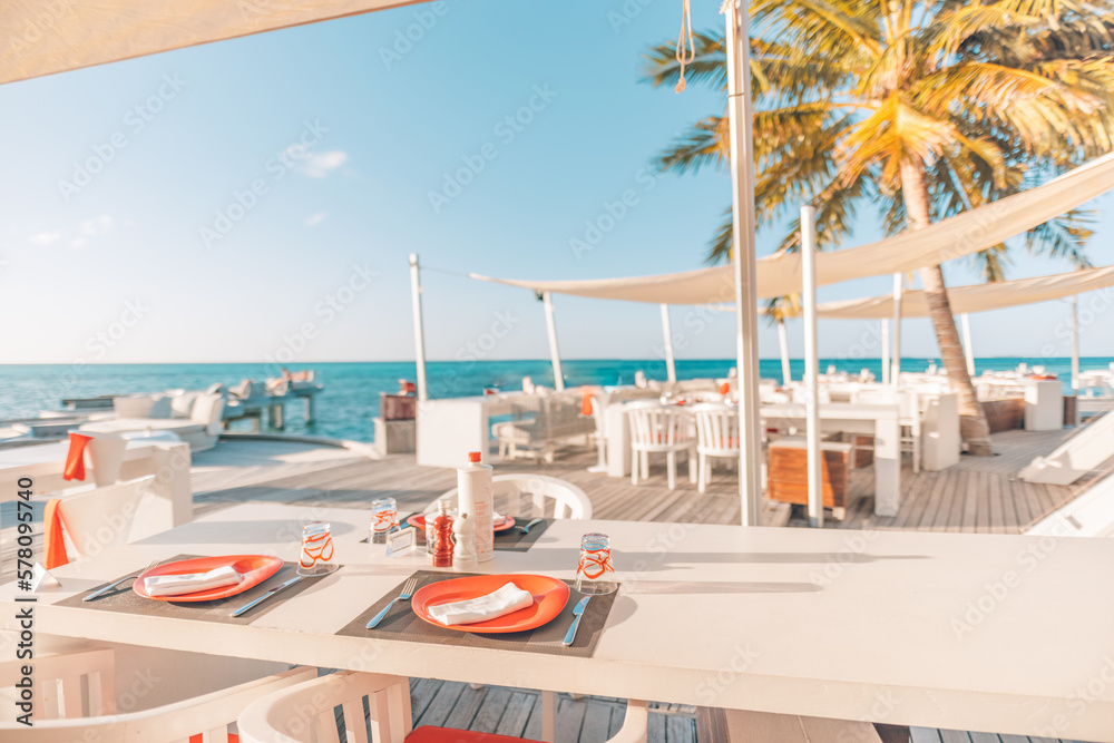 Relaxing outdoor restaurant at the beach. Table setting at tropical beach restaurant. Elegant white wooden tables, chairs under beautiful soft sunset sky, sea view. Luxury hotel or resort restaurant