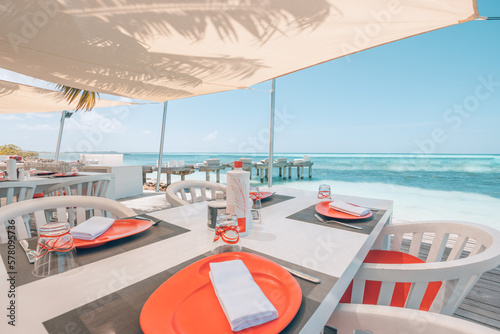 Relaxing outdoor restaurant at the beach. Table setting at tropical beach restaurant. Elegant white wooden tables, chairs under beautiful soft sunset sky, sea view. Luxury hotel or resort restaurant