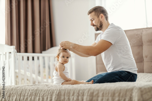 A father is making his daughter's hair while sitting on bed at home.