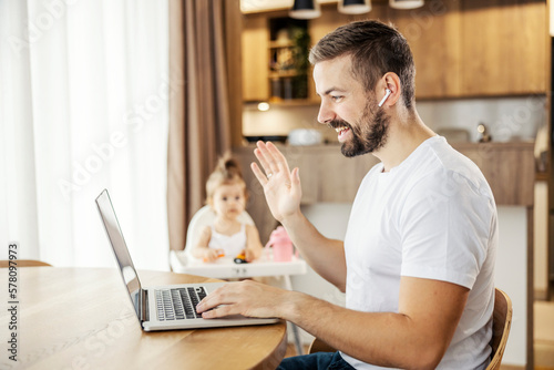 A remote worker is having conference call on a laptop with colleagues and waving while babysitting his girl.