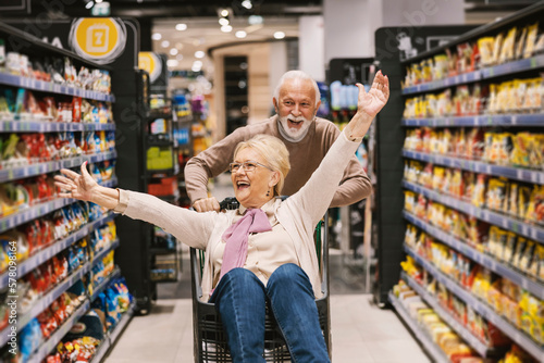 Fotobehang A playful senior couple in love is riding shopping cart at the supermarket and having fun