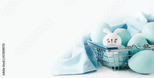 Several blue dyed Easter eggs with cute bunny in metal basket on white table. Easter holiday eggs hunt and painting concept banner.