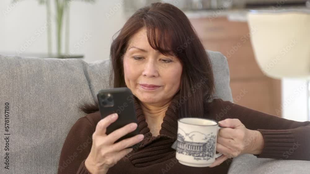 Attractive stylish woman brown sweater enjoying texting and checking email on mobile phone drinking morning coffee sitting on sofa in home living room. Beautiful asian female holding hot cup.