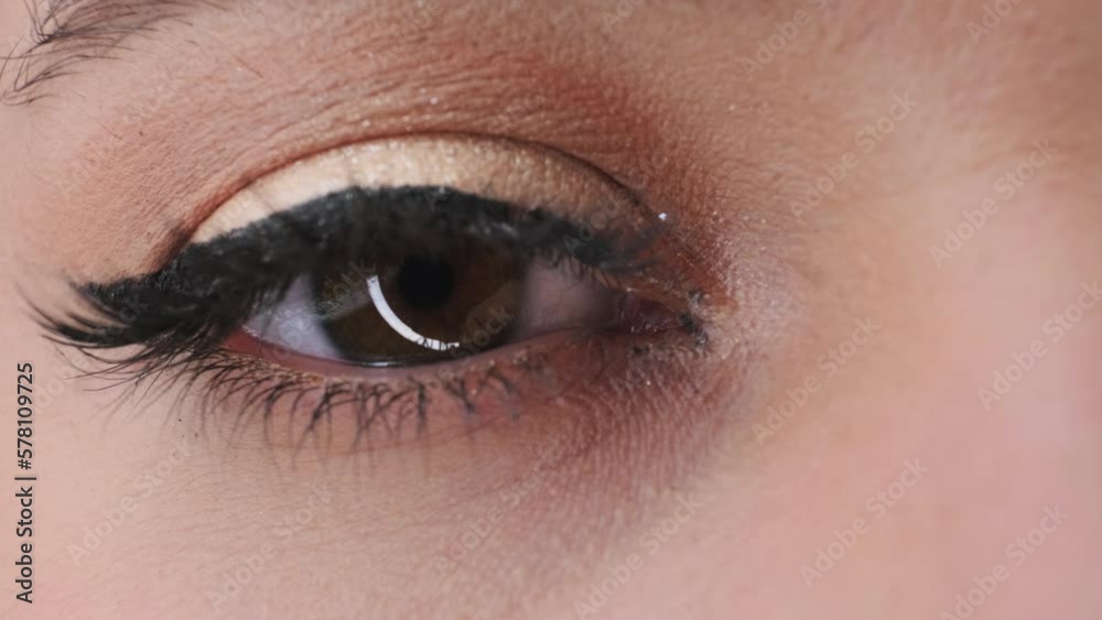Eye of young woman with dark arrow and long artificial lashes. Female person with bright reflection of light on brown iris blinks fast extreme closeup Eye of young woman with dark arrow and long artificial lashes. Female person with bright reflection of light on brown iris blinks fast extreme closeup