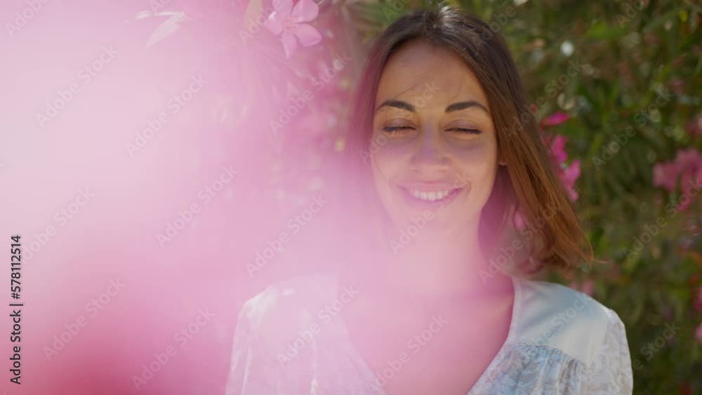 Close up face of pretty brunette tanned woman standing at pink flowers and looking at camera with charming smile. Concept of beauty and perfume Close up face of pretty brunette tanned woman standing at pink flowers and looking at camera with charming smile. Concept of beauty and perfume