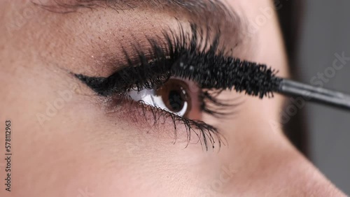 Young woman with brown eyes applies black mascara on long eyelashes. Female paints artificial lashes with brush on blurred background macro