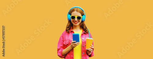 Portrait of stylish modern happy smiling young woman in headphones listening to music with smartphone wearing pink jacket on orange background © rohappy