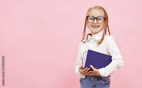 Adorable schoolgirl with funny pigtails on head holding textbooks in hands smiling in camera and fixing glasses finger on pink background, prepares for exam. Back to School. Childhood and education