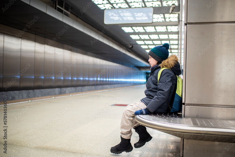 a smiling boy in a winter jacket and hat is waiting for an approaching train on a subway platform