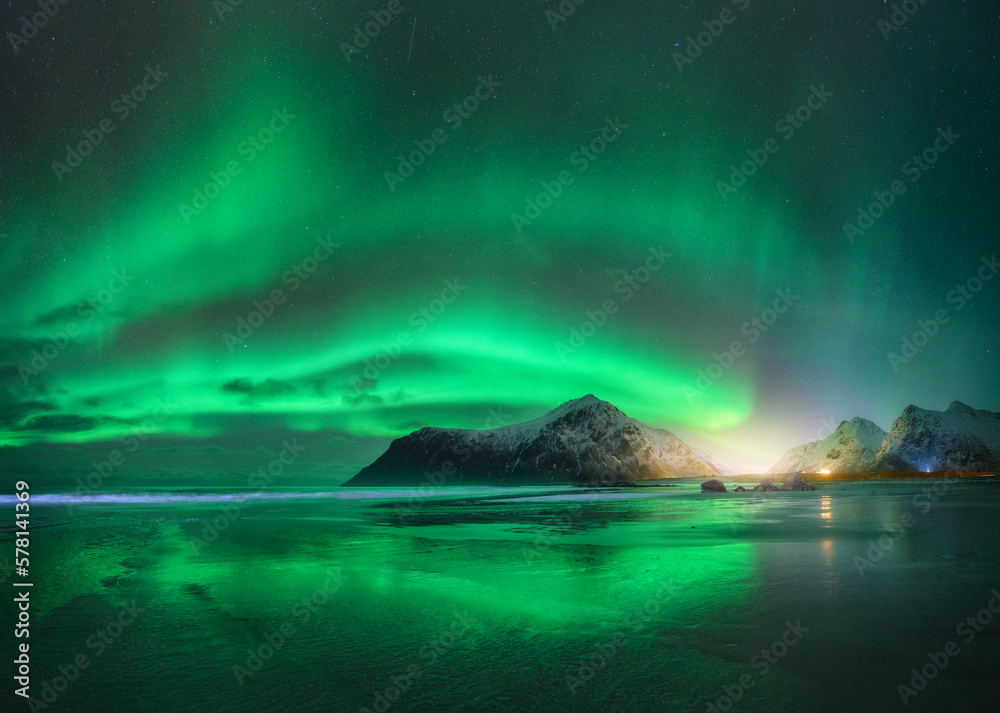 Northern Lights and beach in Lofoten islands, Norway. Beautiful Aurora borealis. Starry sky with polar lights. Night winter landscape with aurora, sea with sky reflection in water, snowy mountains
