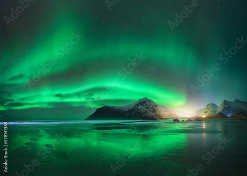 Northern Lights and beach in Lofoten islands, Norway. Beautiful Aurora borealis. Starry sky with polar lights. Night winter landscape with aurora, sea with sky reflection in water, snowy mountains