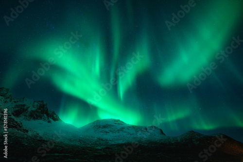 Northern lights over the snowy mountains at night in Lofoten, Norway. Aurora borealis above the snow covered rocks. Winter landscape with polar lights, mountain peak. Starry sky with bright aurora