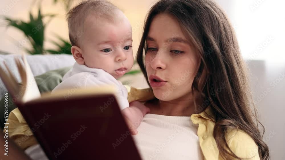 Portrait of cute young family spend leisure time together when mother reading book literature to her little baby soothes the child with his pleasant voice laying on sofa at light room Portrait of cute young family spend leisure time together when mother reading book literature to her little baby soothes the child with his pleasant voice laying on sofa at light room