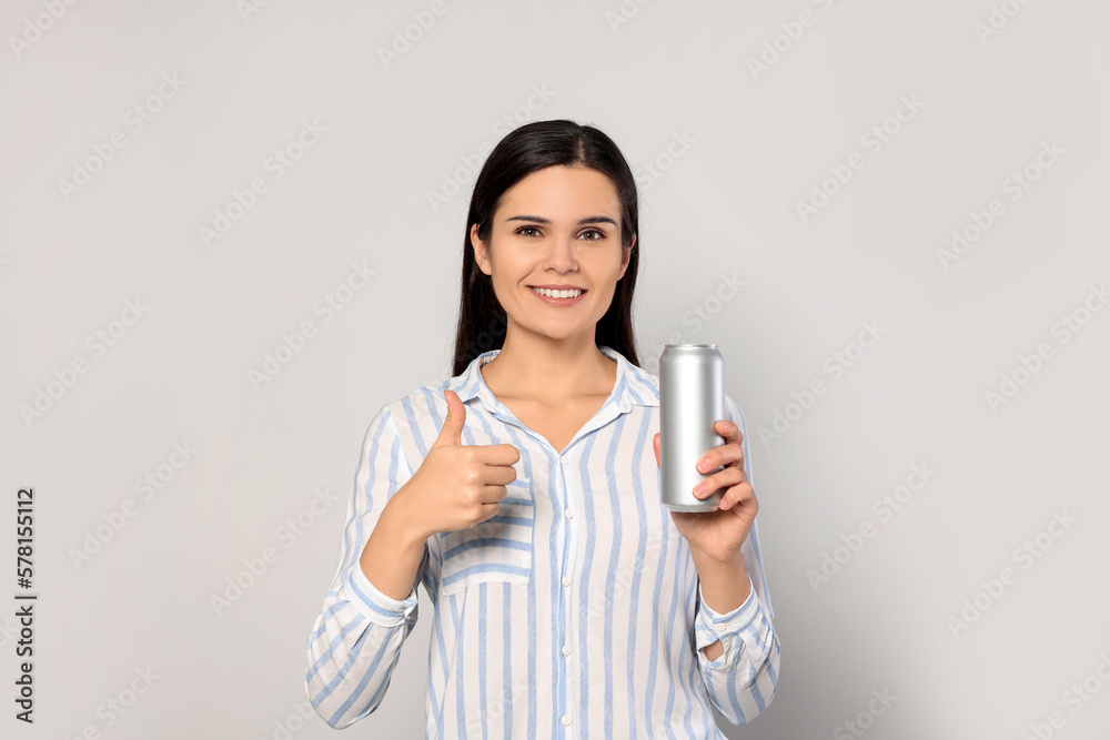 Beautiful young woman holding tin can with beverage and showing thumb up on light grey background Beautiful young woman holding tin can with beverage and showing thumb up on light grey background