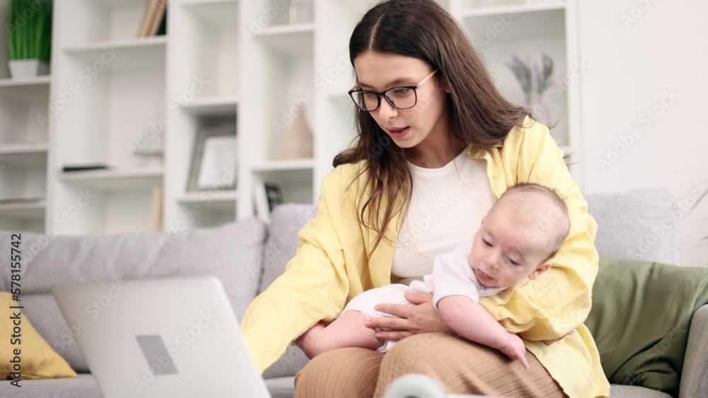 Portrait of young business mother with glasses hold her baby while working on laptop computer planning day or details of new project at light home office Successful woman balancing work and motherhood Portrait of young business mother with glasses hold her baby while working on laptop computer planning day or details of new project at light home office Successful woman balancing work and motherhood