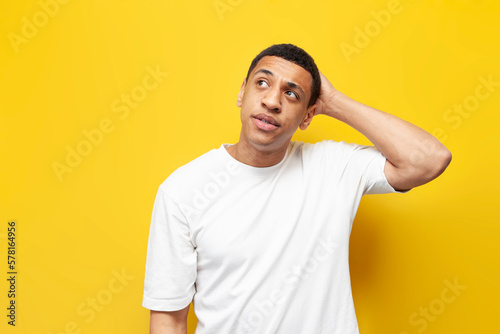 young pensive guy afro american confused scratches his head and remembers on yellow isolated background