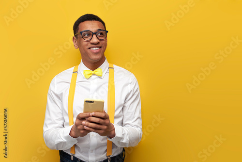 young pensive afro american guy in festive outfit with bow tie and suspenders uses smartphone on yellow background