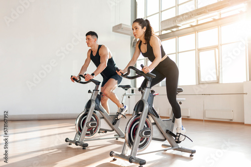 athletic couple of cyclists train on static bicycle simulator, man trainer and fitness woman go in for sports together