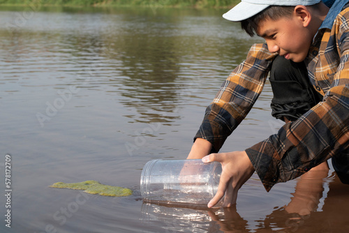 Asian schoolboy holding transparent plastic tube to keep freshwater algae from local river inside to study the fertility of the river's nature and to do environment school project work, in motion.