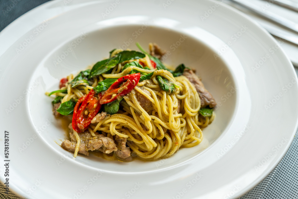 Delicious Thai fusion food, stirred fried spaghetti with thai green curry and meat. Beautiful Thai food in modern white dish on a table in restaurant, natural light from window.