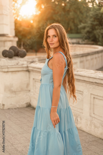 Woman sunset blue dress. Portrait of a woman with long hair and a blue dress against the backdrop of the setting sun and a white building. Lifestyle, walking around the city © svetograph