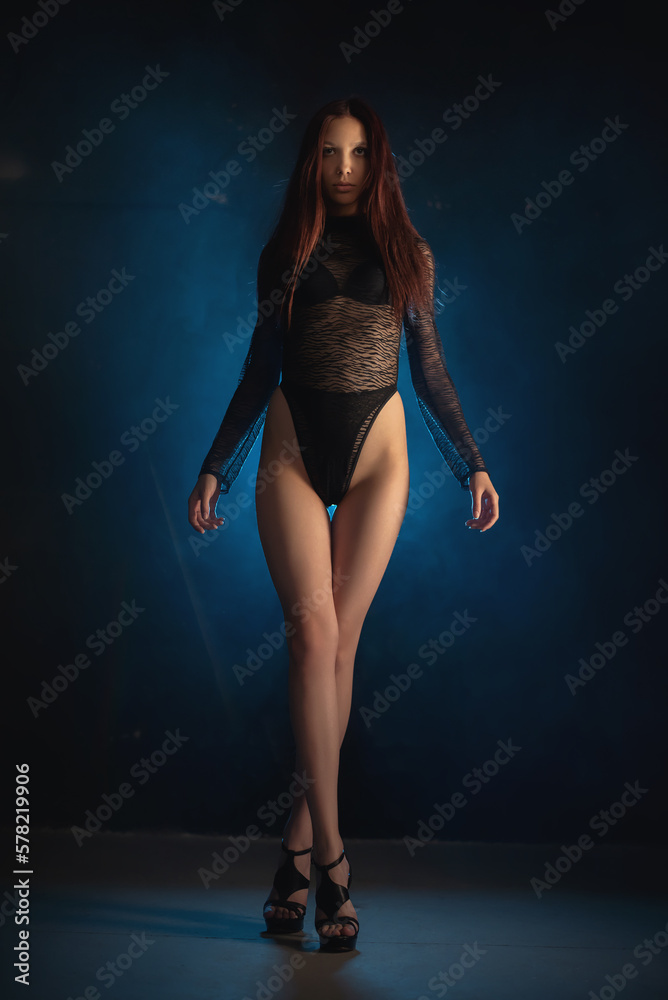 Obraz premium Beautiful girl a dancer in the black body suit on the dark background in the blue lights.