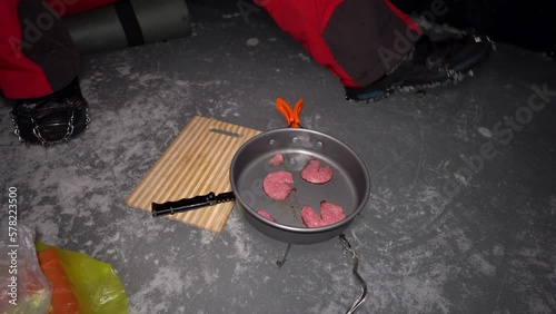 A guy cooks food on gas inside an ice cave. The climber lights the burner, cuts sausage and tomato. Breaks the eggs and fry in a frying pan. Steam from food. The ice wall is blue and the floor is gray photo