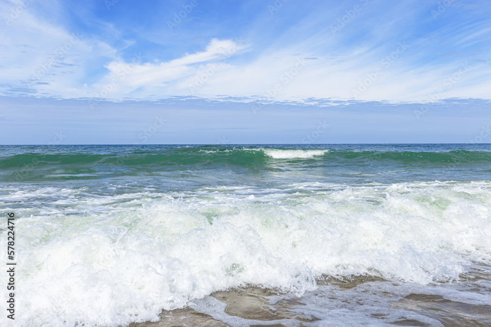 Blue cloudy sky above blue sea water, waves crashing sandy beach, windy weather. Summer vacation and holiday travel concept. Seascape nature background, beautiful seashore with water foam