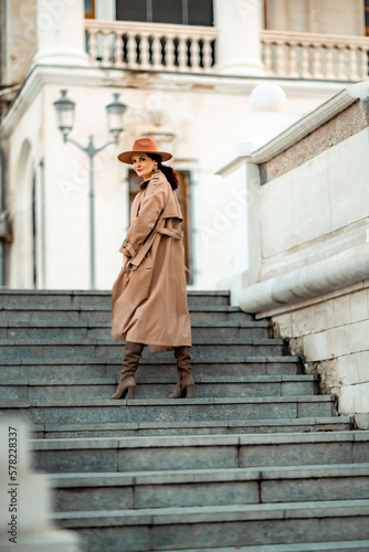 Outdoor fashion portrait of young elegant fashionable brunette woman, model in stylish hat, choker and light raincoat posing at sunset in European city.