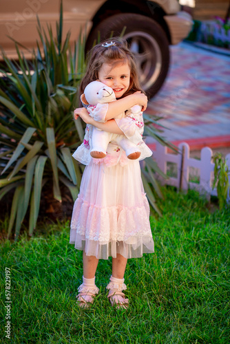Cute little girl is holding a plush toy, playing in the yard. Beautiful little girl smiles in a smart dress.